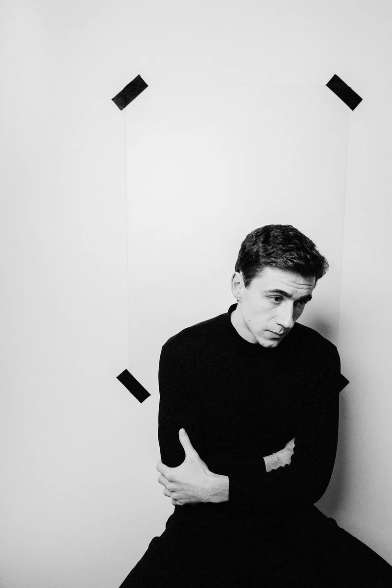Man in a black turtleneck sitting by a white wall with tape marks, looking down