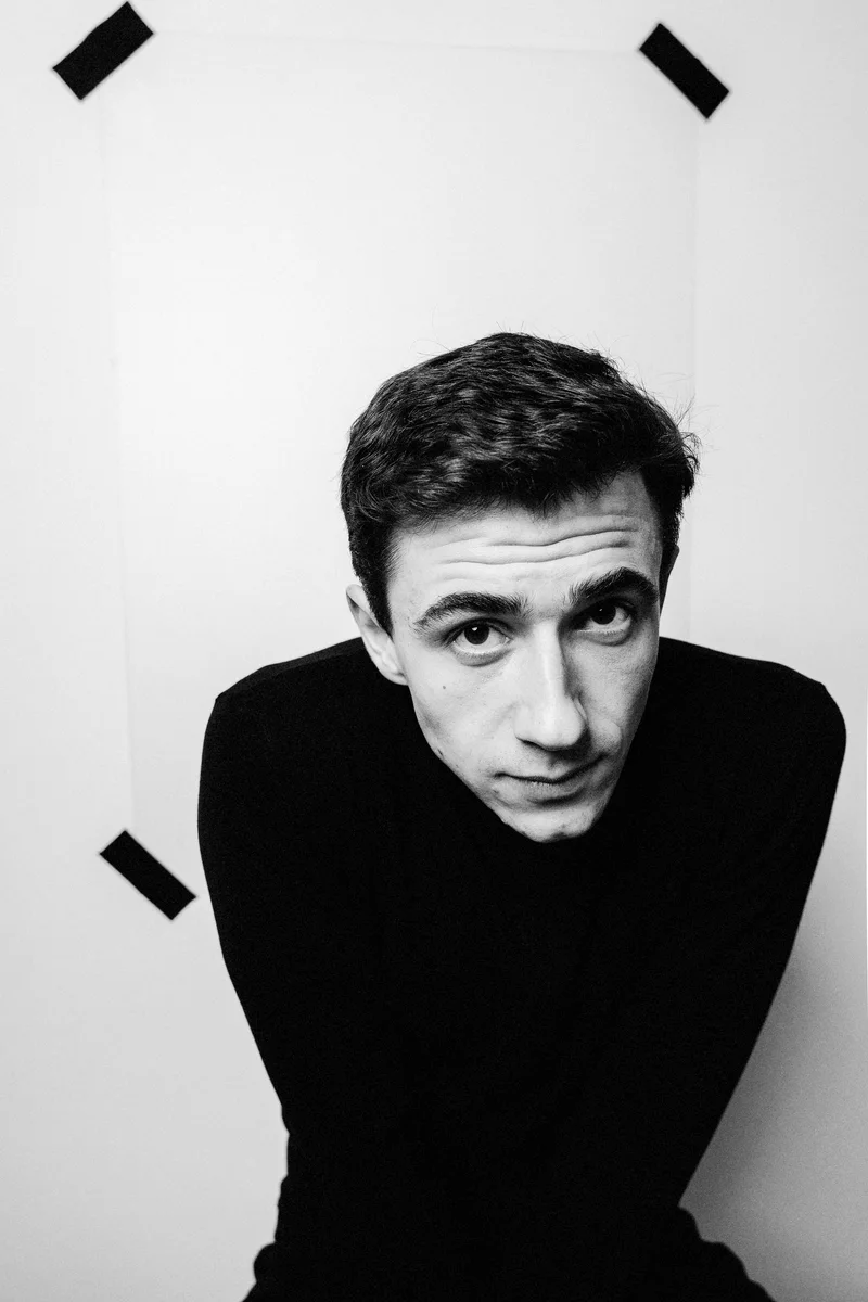 Man in a black turtleneck leaning towards the camera against a white wall