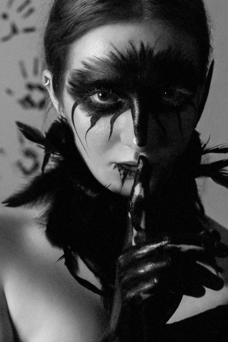 Woman with black feathered make-up around one eye and a finger to her lips