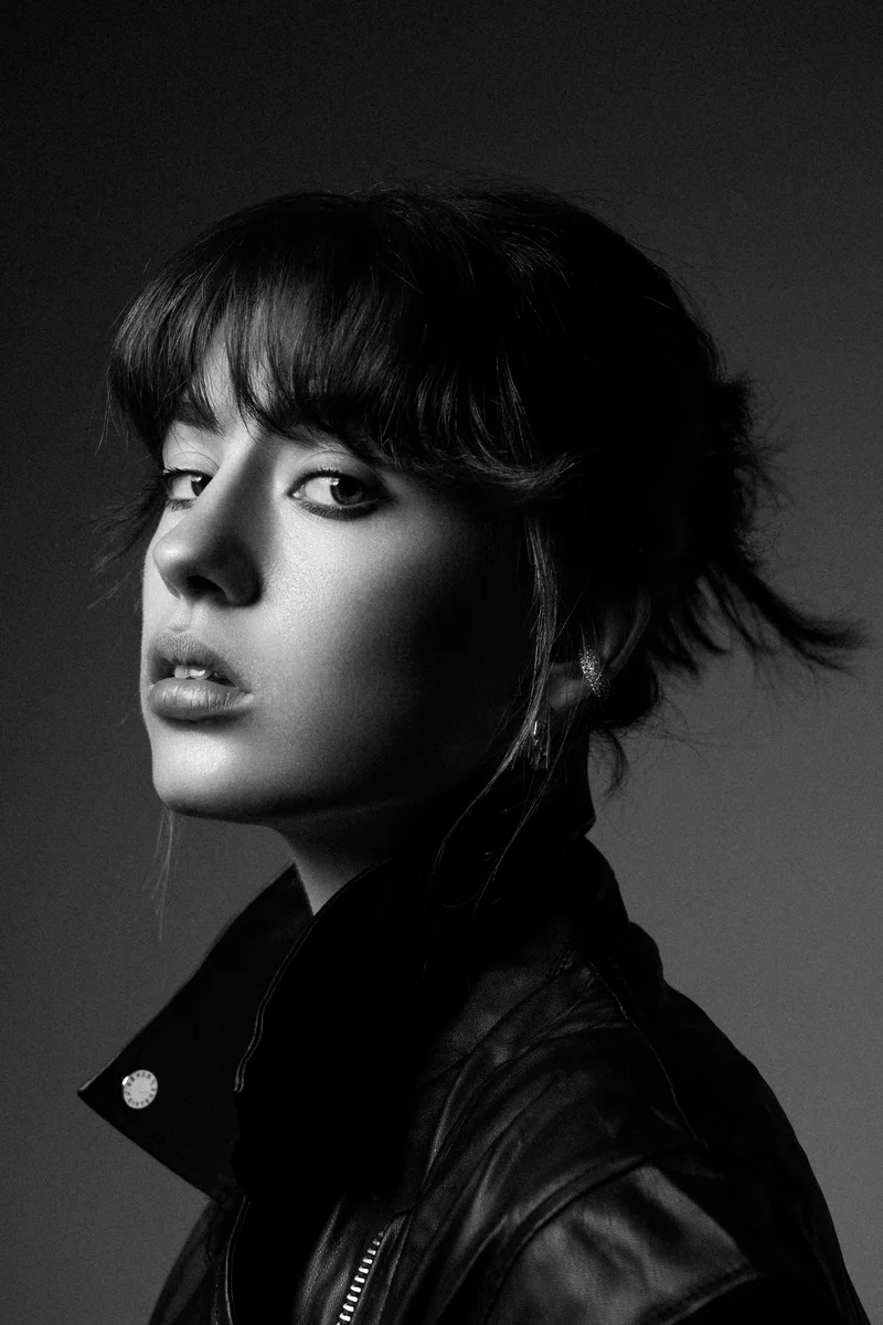 Woman with a short fringe in a leather jacket glancing over her shoulder against grey