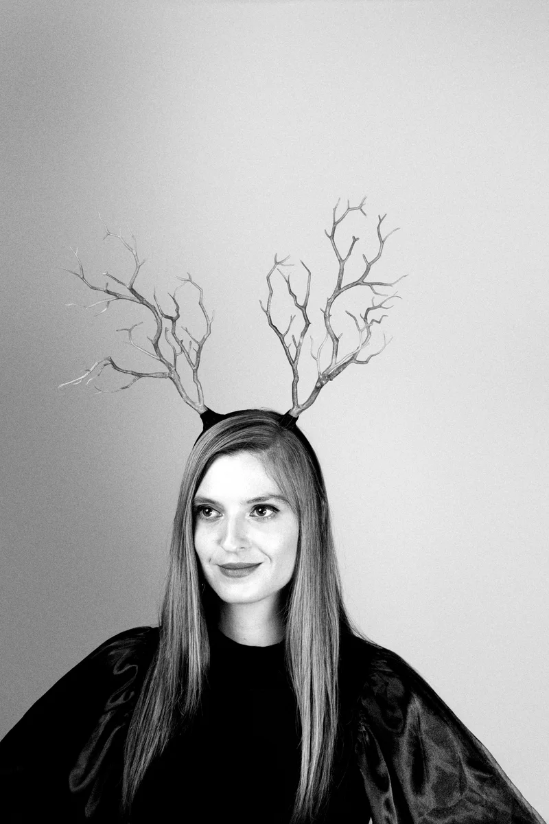 Smiling woman with branch-like antlers on her head and a black cape against a pale background