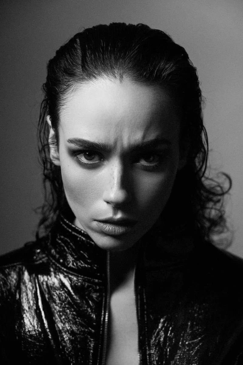 Woman with wet slicked-back hair in a glossy black jacket, with an intense gaze