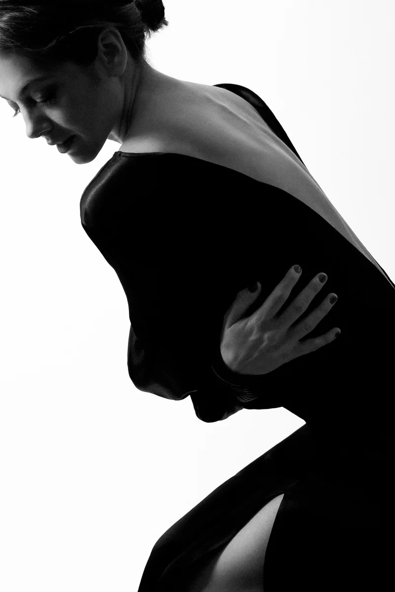 Woman in a black open-back dress bending forward against white