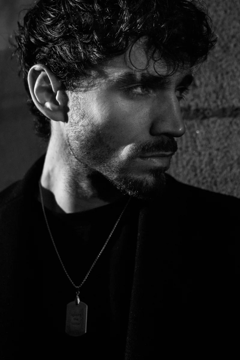 Man with curly hair and a beard wearing a pendant against a stone wall
