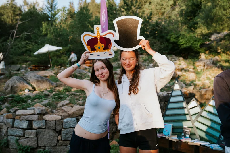Two women posing with crown and top-hat props at an outdoor event
