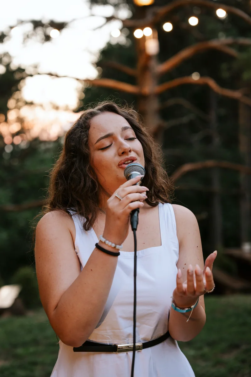 Woman singing into a microphone outdoors beneath string lights