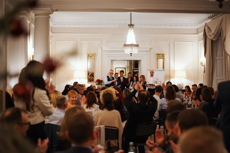 Guests at tables applauding in a white hall with a chandelier