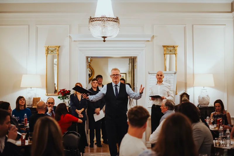 Host with outstretched arms speaking between the guests' tables