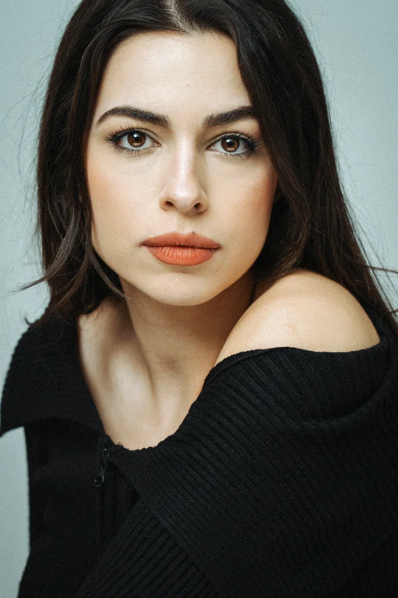 Dark-haired woman in a black jumper slipping off one shoulder against light grey
