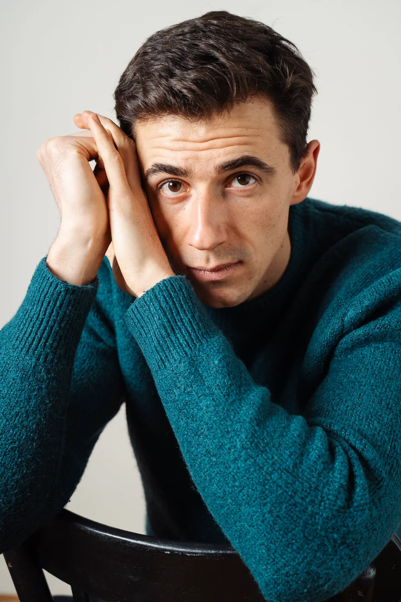 Man in a teal sweater with his palms pressed together beside his face against a light background