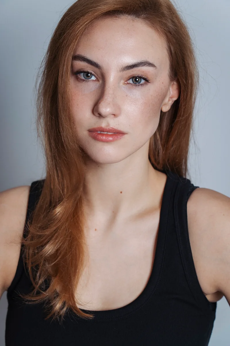 Red-haired woman in a black vest looking ahead against a grey studio backdrop