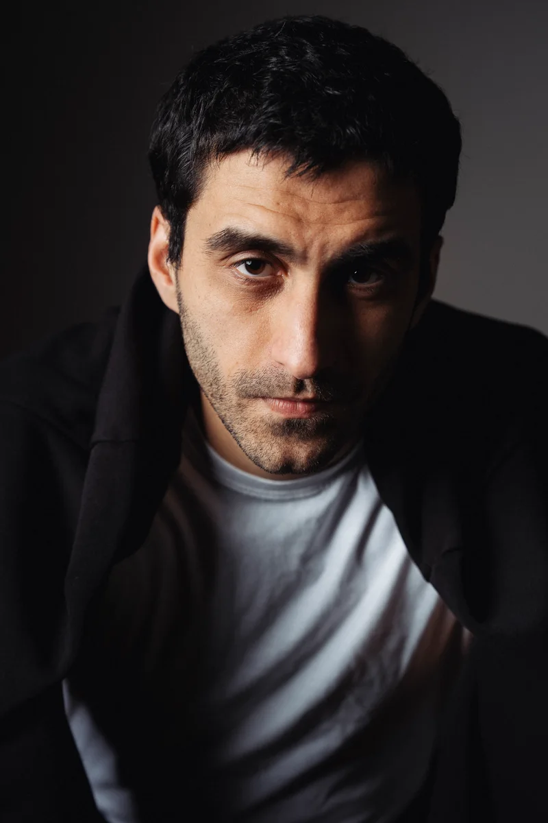 Man with stubble in a black jacket and a white T-shirt against a dark background