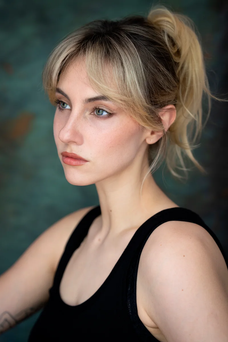 Blonde woman with her hair tied up in a black top looking aside against teal