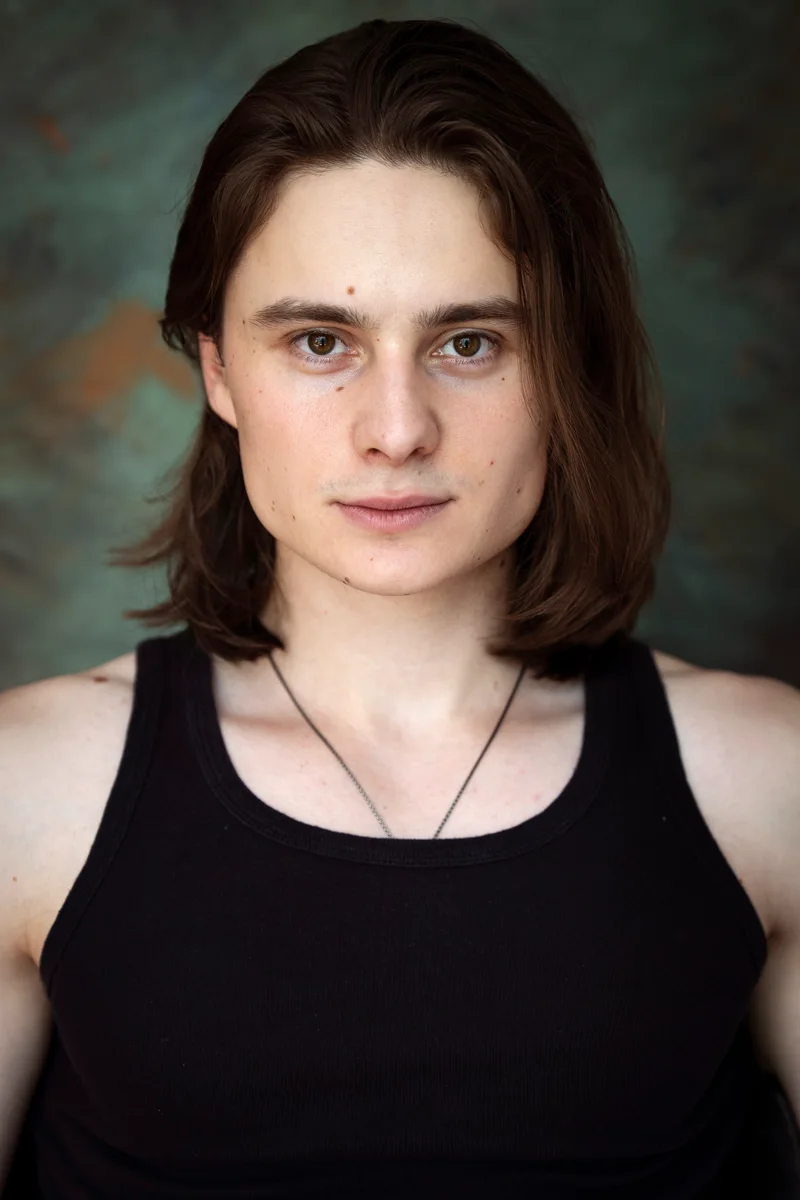 Person with shoulder-length hair in a black tank top and a pendant against teal