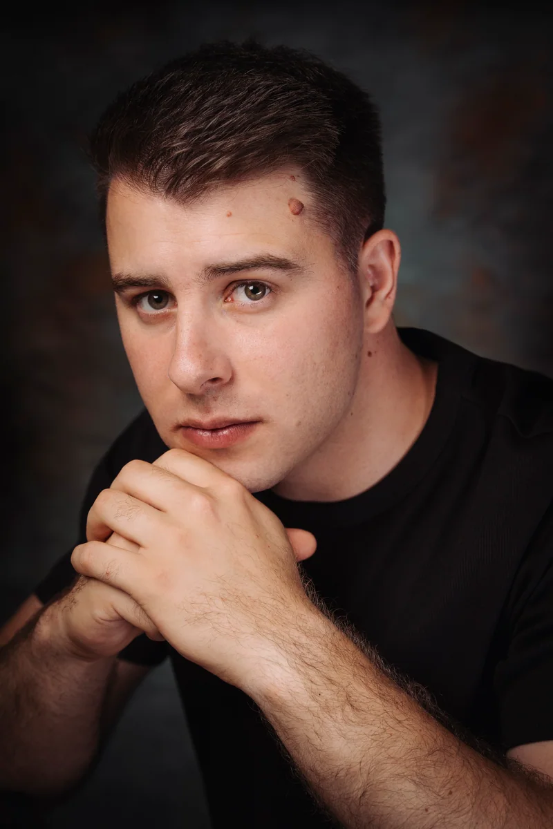 Man with short dark hair in a black T-shirt, hands clasped under his chin