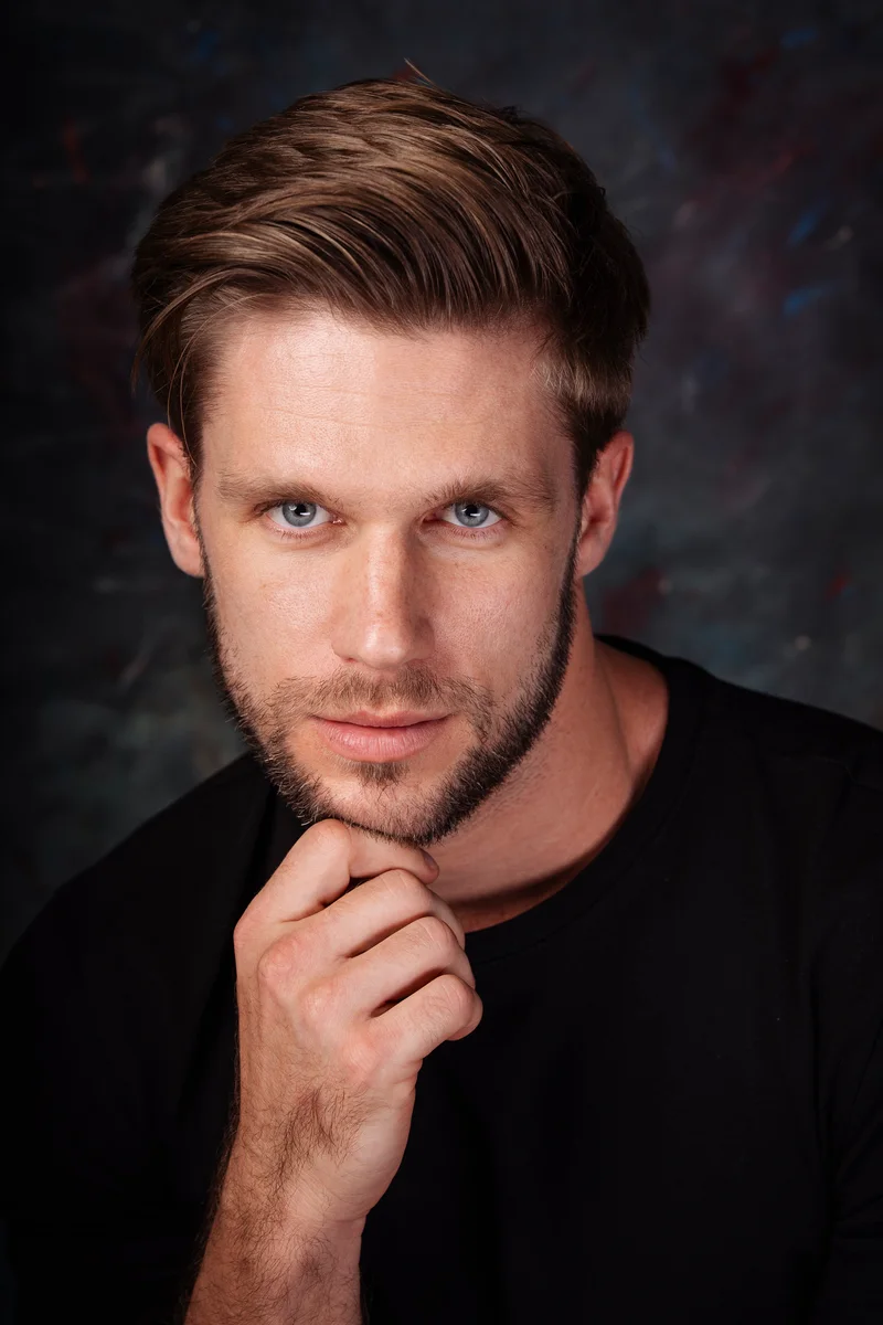 Man with a short beard and black T-shirt holding his hand beneath his chin
