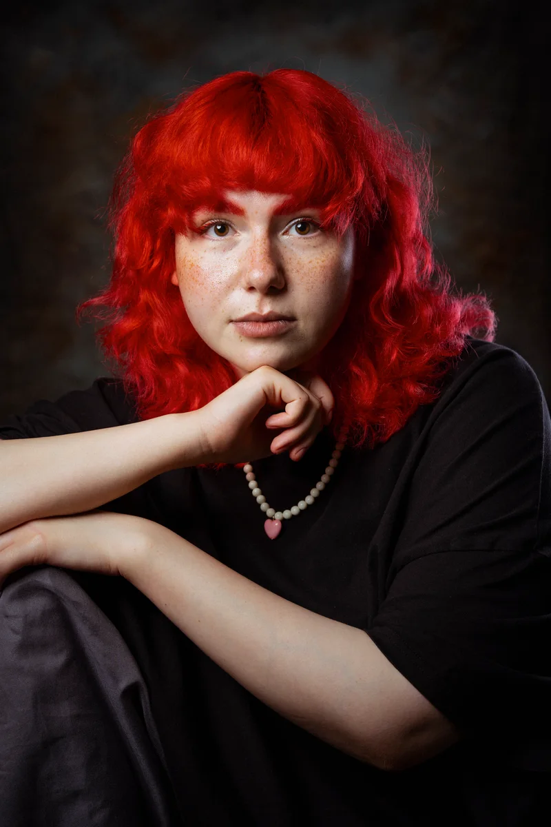 Woman with bright red curly hair and a pearl necklace with a heart, her hand under her chin