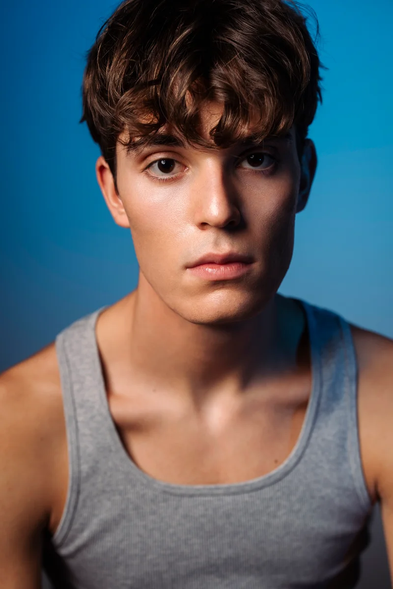 Frontal portrait with short curly hair and a grey vest against a blue backdrop
