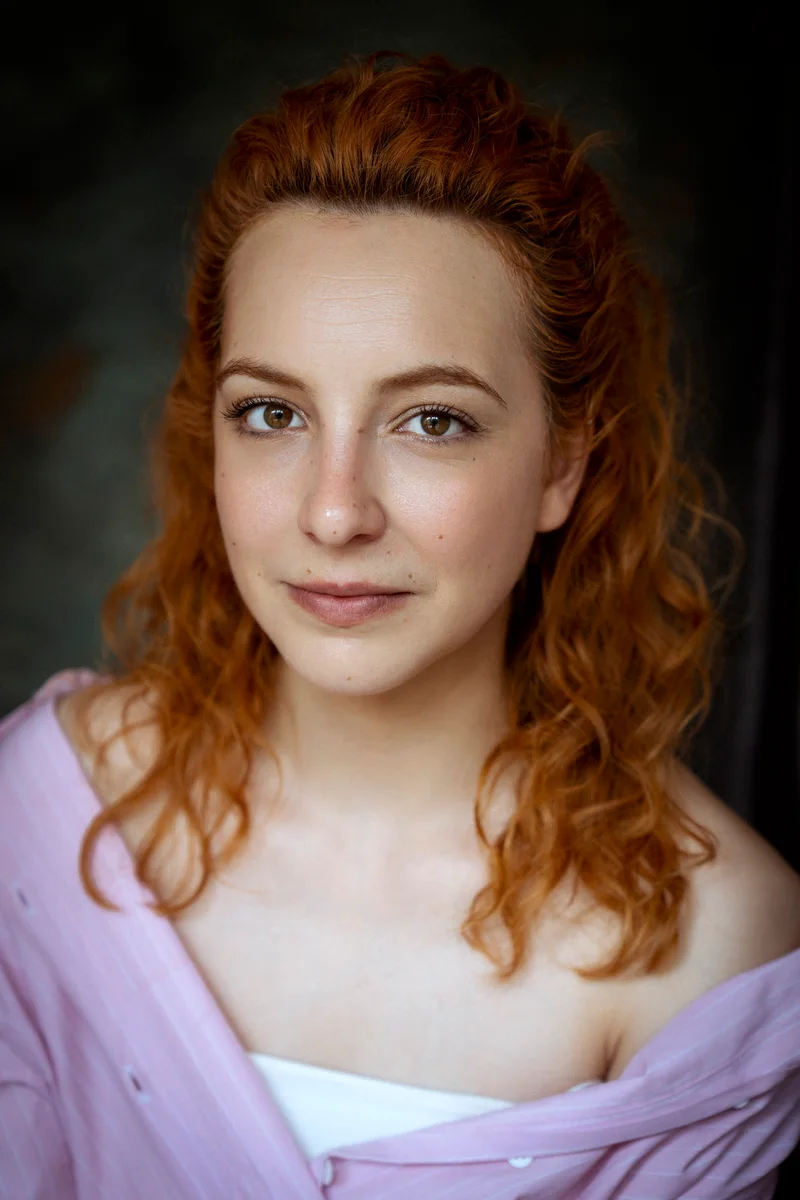 Red-haired woman with swept-back hair, a white top and purple drapery