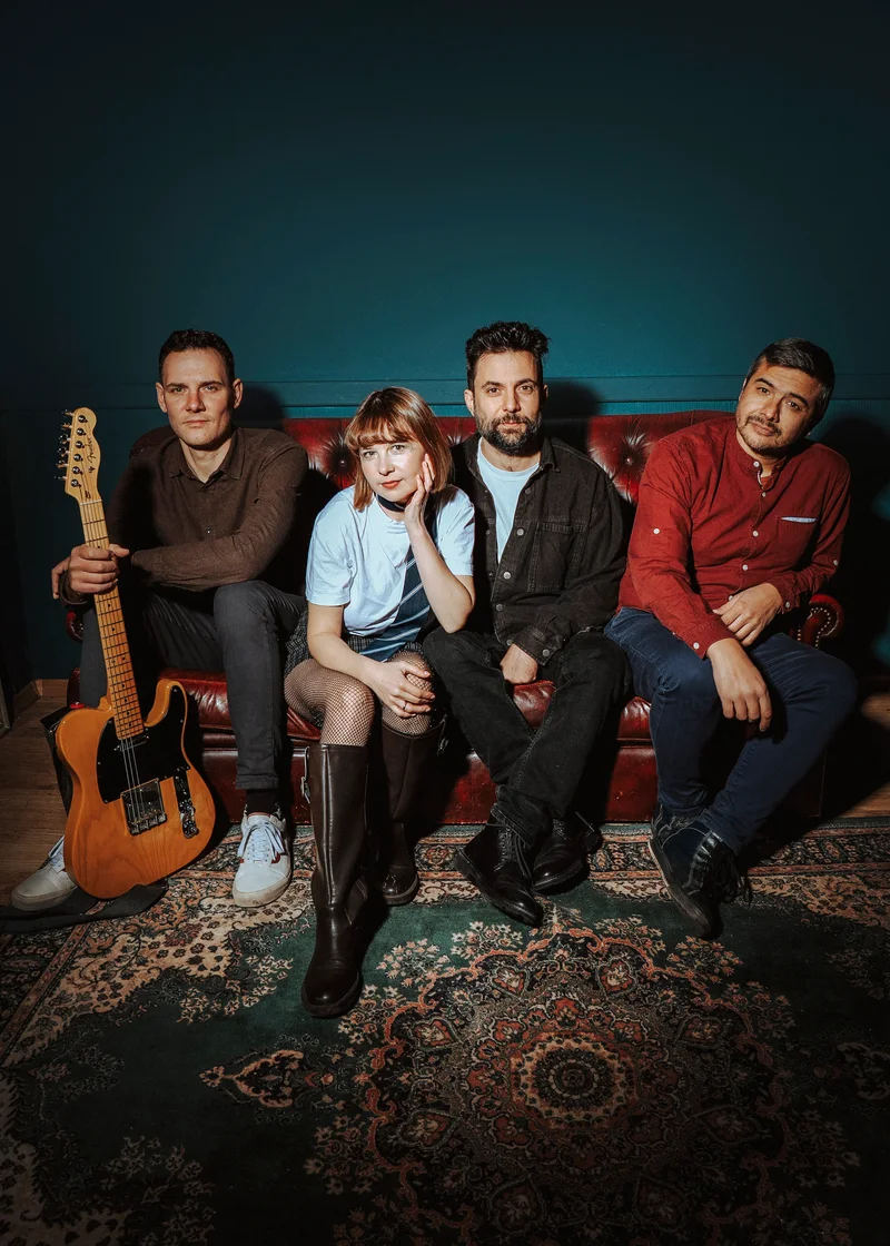 Four-piece band on a red leather sofa against a dark teal wall