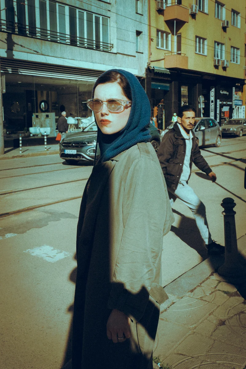 Woman in a blue headscarf and sunglasses on a street as a man crosses behind her