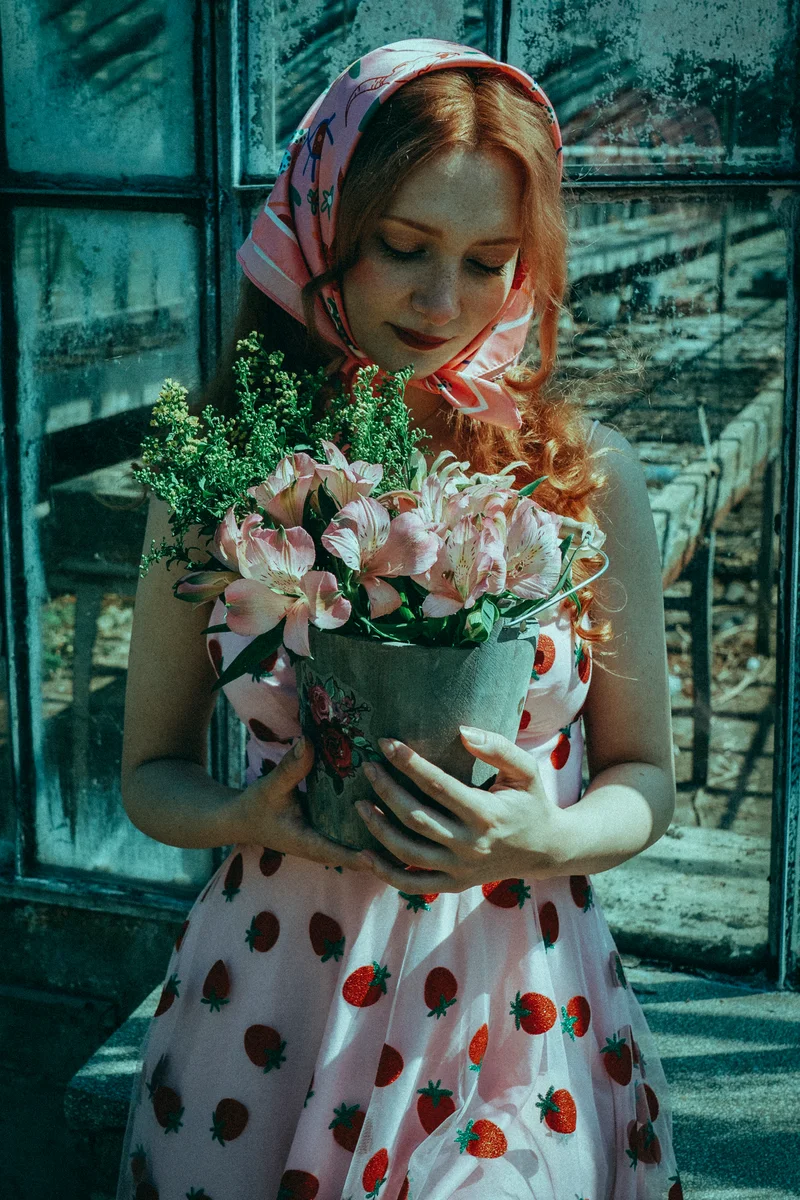Red-haired woman in a headscarf and polka-dot dress holding flowers beside an old door