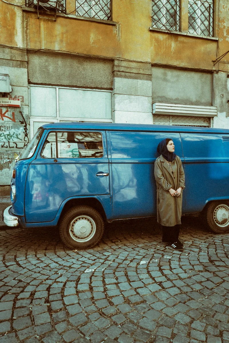 Woman in a grey coat leaning on a blue vintage van on a cobbled street