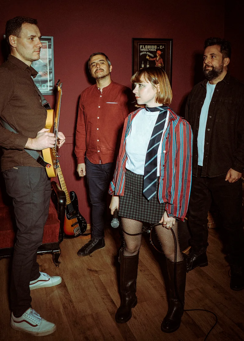 Four-piece band in a red room, the woman in a tie at the centre