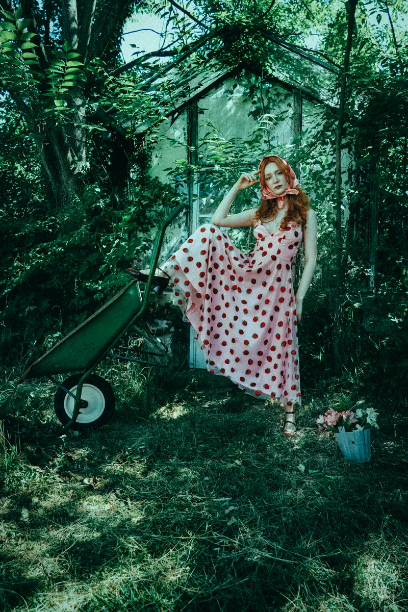 Woman in a polka-dot dress sitting on the edge of a green wheelbarrow among trees