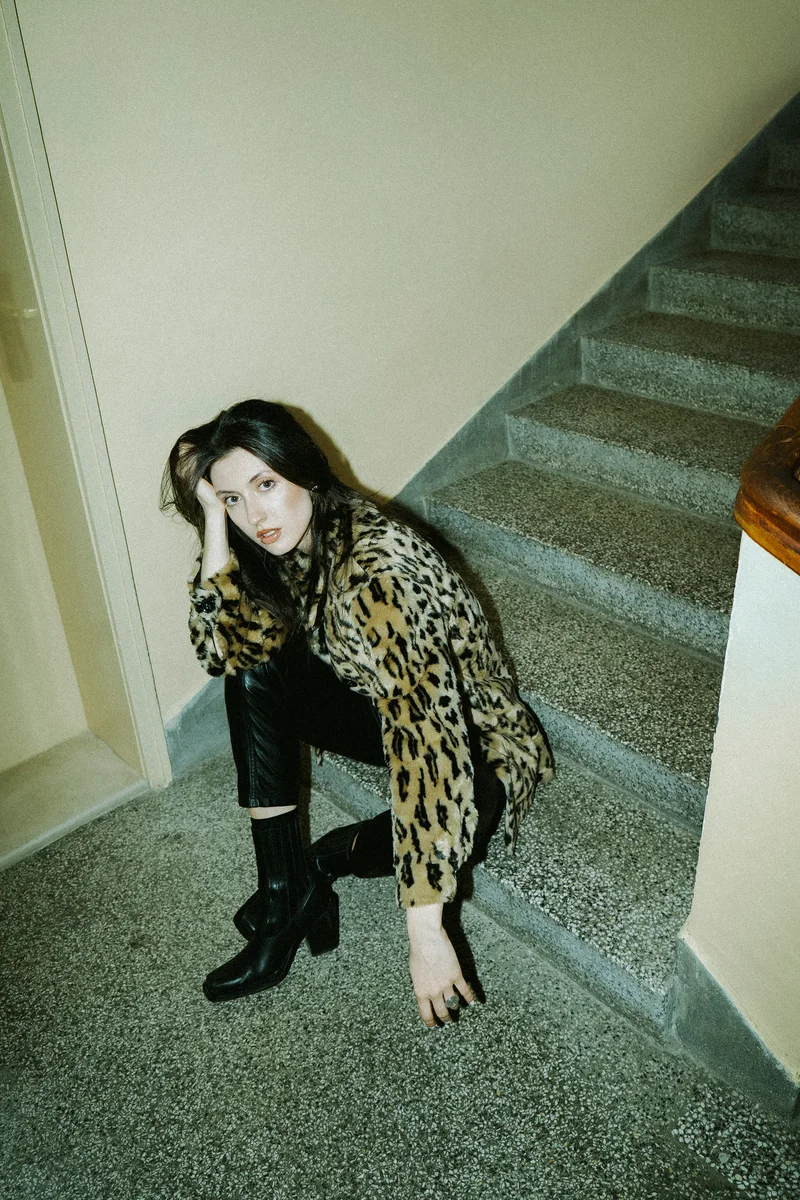 Woman in a leopard-print coat crouching on stairs in an indoor hallway