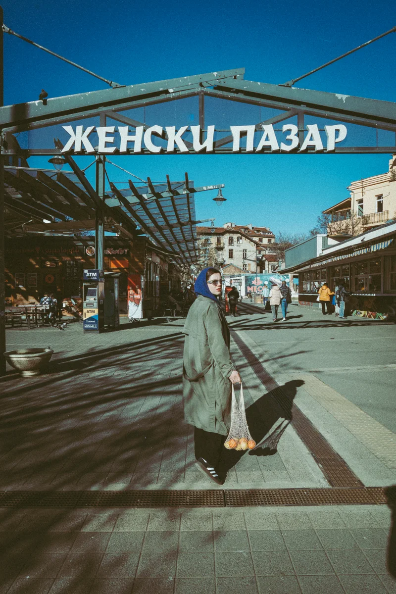 Woman with a headscarf carrying a string bag of oranges beneath the Zhenski Pazar market sign