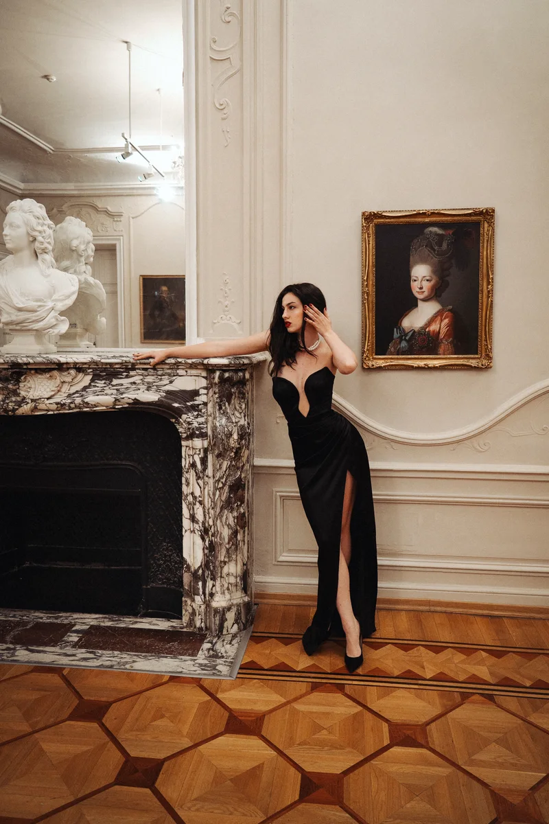 Woman in a black evening gown leaning on a marble fireplace beneath a portrait