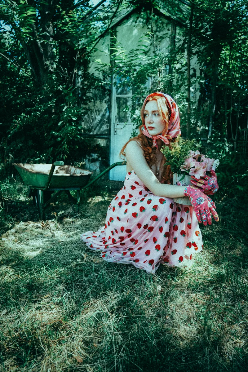 Woman with a red headscarf and polka-dot dress sitting with flowers in a garden