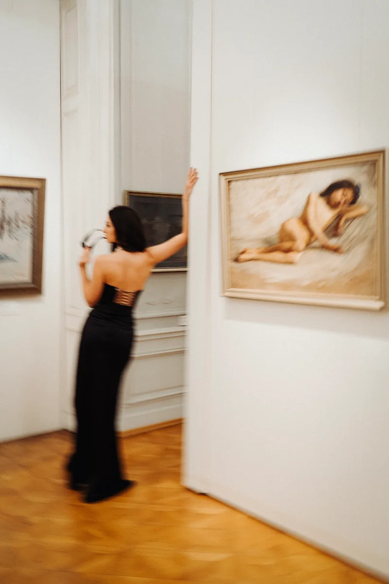 Woman in a black gown viewing a painting in a gallery, her back to the camera