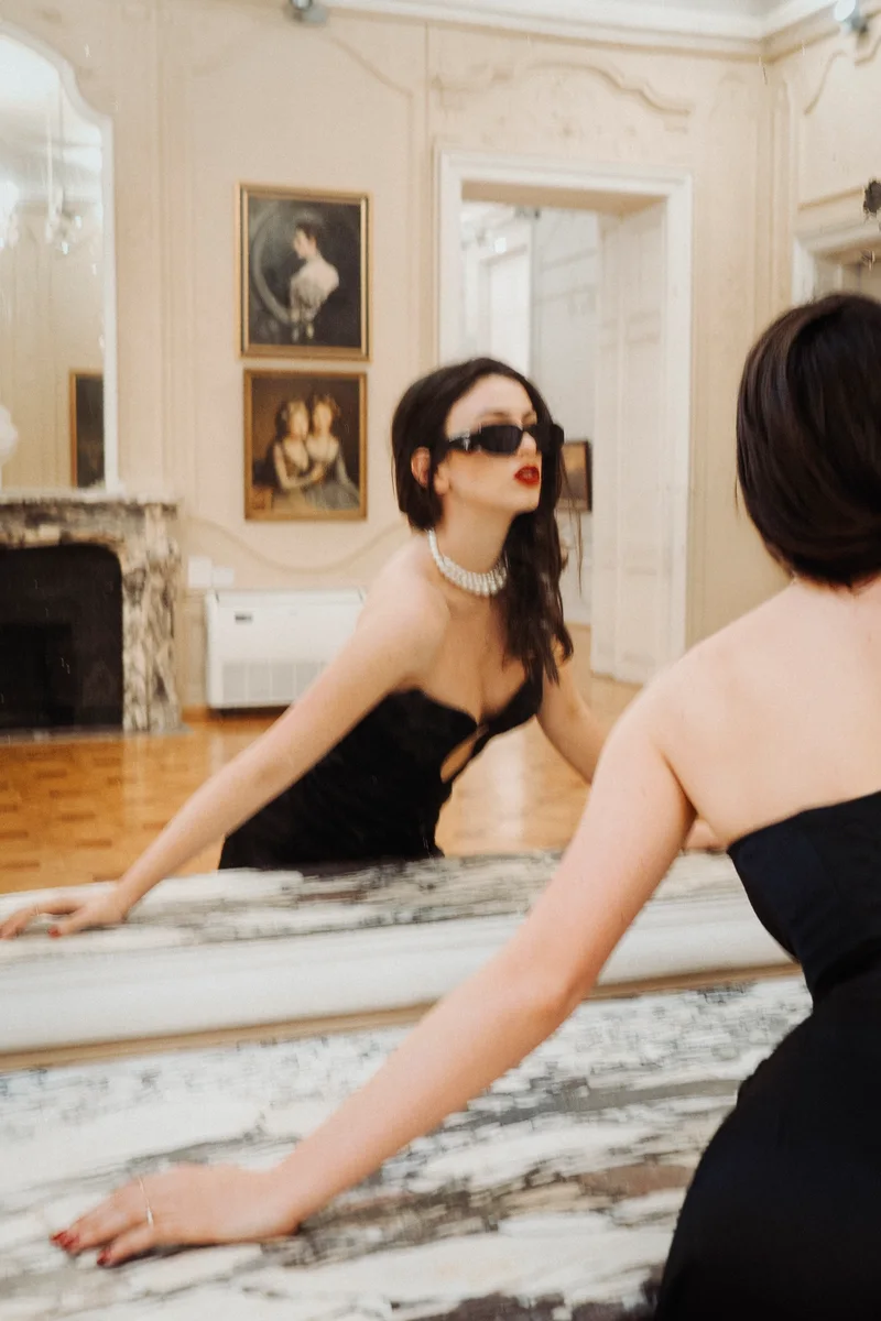 Woman in sunglasses leaning on marble beside her reflection in a mirror