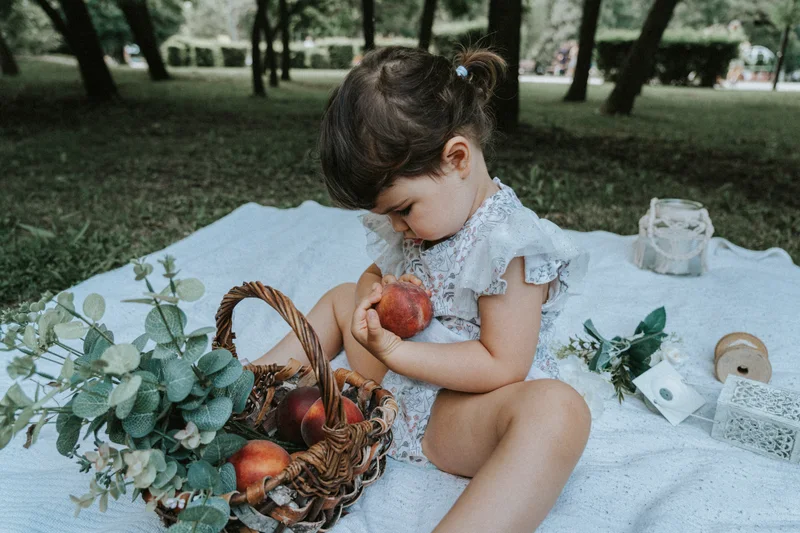 Small child on a white blanket in the park looking at a piece of fruit