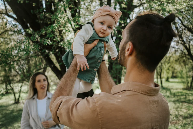 Man lifting a baby with a pink bow in the park as a woman looks on