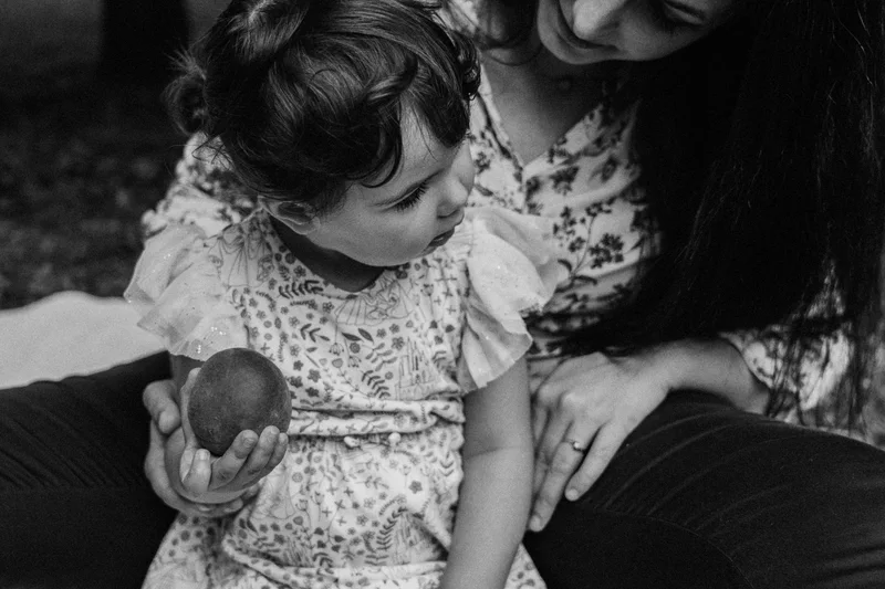 Child holding a piece of fruit on a woman's lap, in black and white