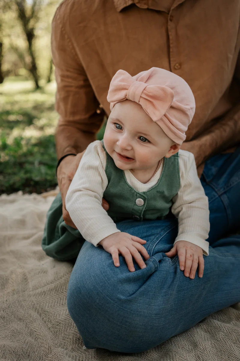 Smiling baby in a pink bow hat sitting in front of a man on a blanket