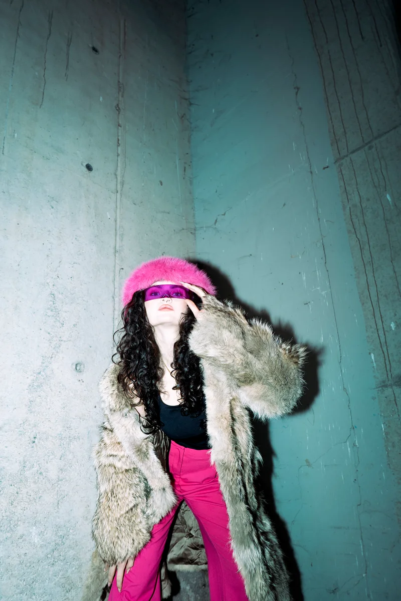 Woman in a pink hat and fur coat holding her hands beside her face against a concrete wall