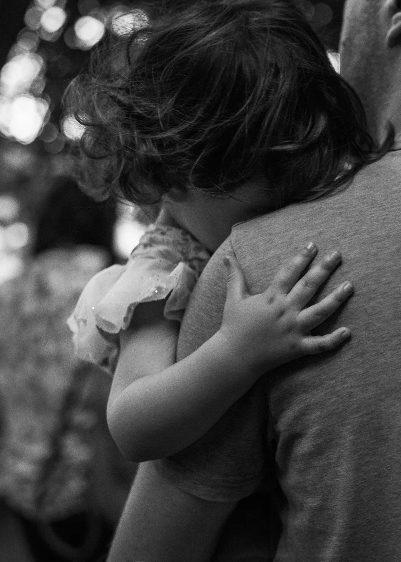 Child with curly hair resting their head on an adult's shoulder, in black and white