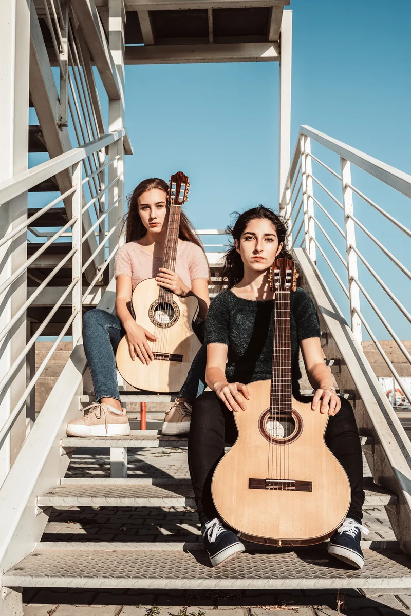 Two women posing with guitars held upright on a white staircase under a blue sky