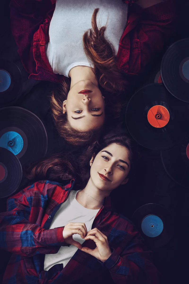 Two women lying head to head among vinyl records, one making a heart with her hands