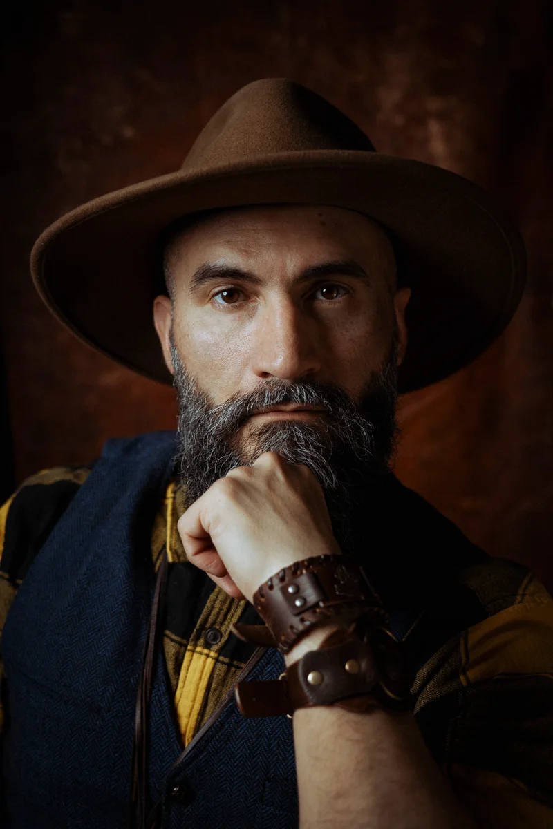 Man with a greying beard and a brown hat, resting his chin on his fist