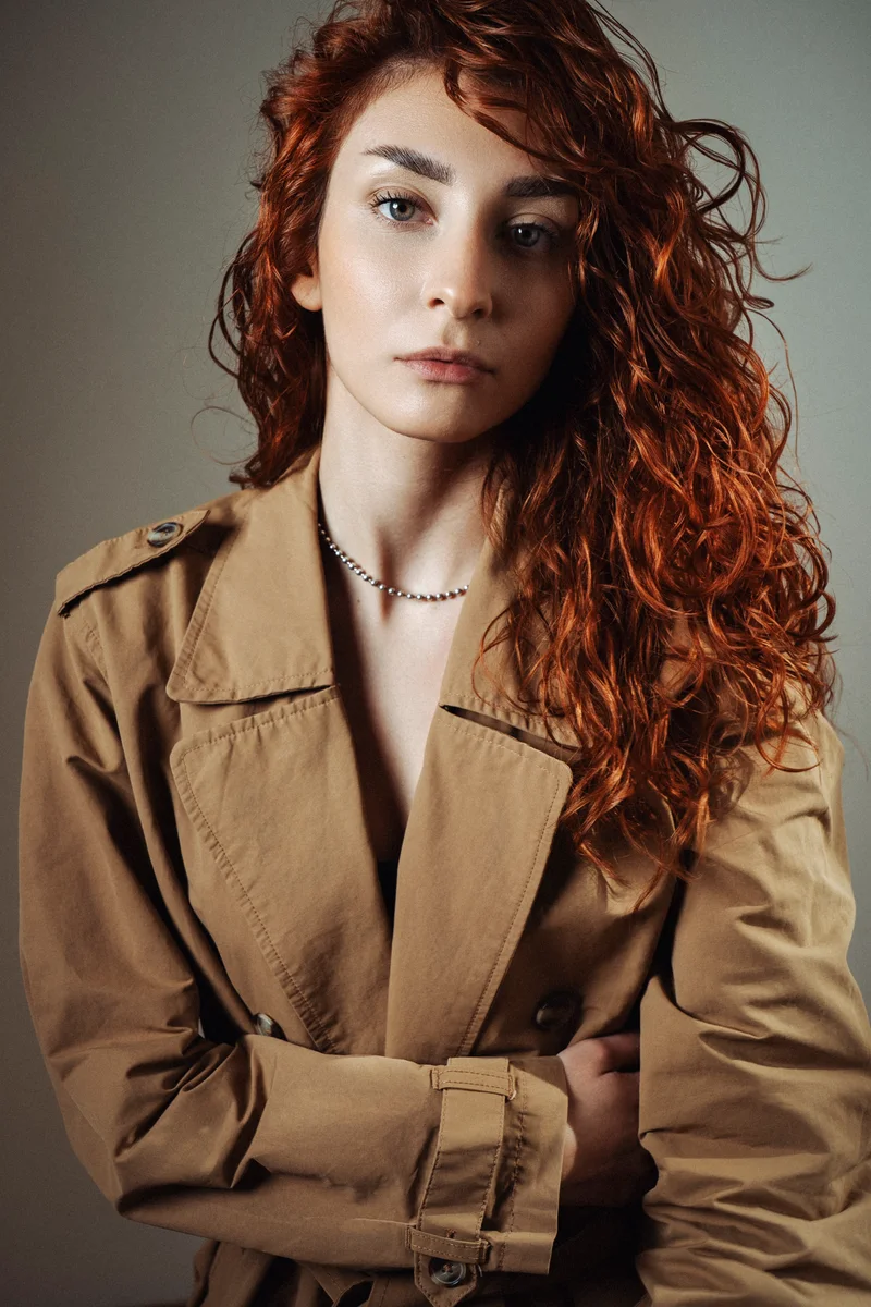 Curly red-haired woman in a beige trench coat with her arms crossed