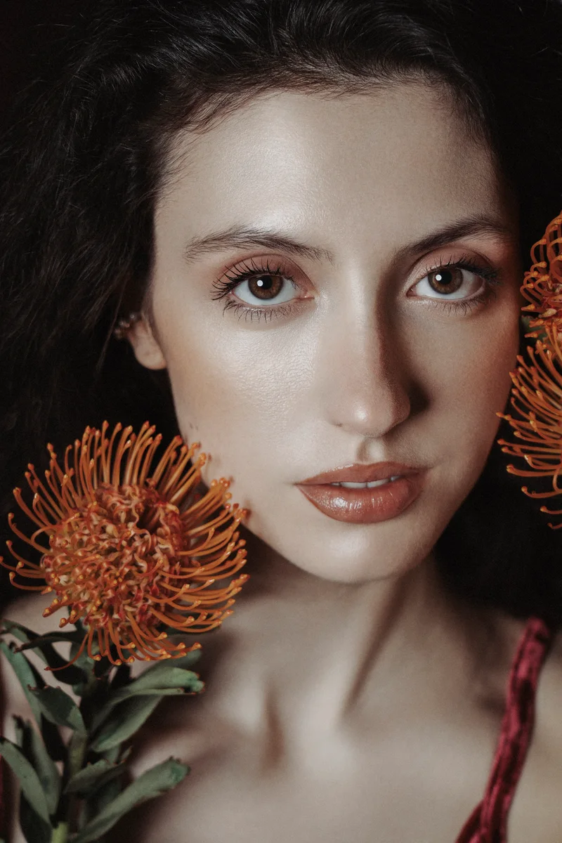 Woman with swept-back hair and an orange flower beside her face in soft light