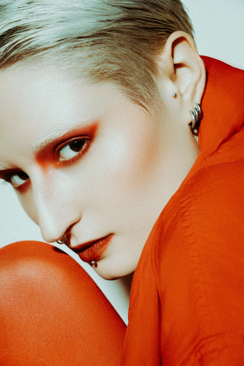 Woman with short blonde hair in an orange sweater leaning towards her shoulder