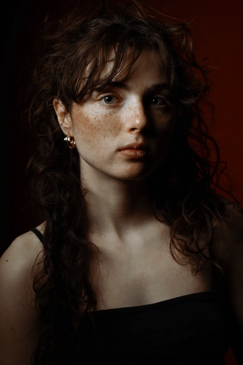 Freckled woman with curly brown hair against a dark red background