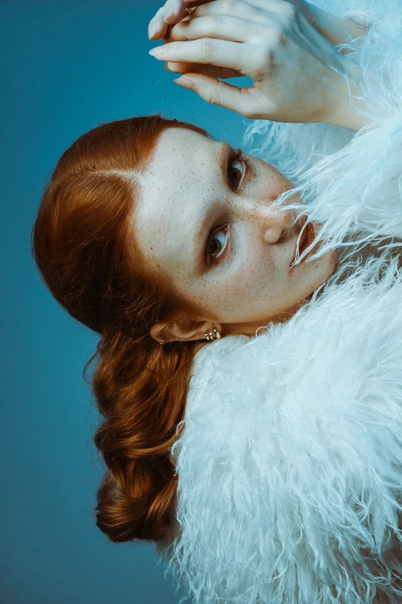 Red-haired woman reclining in white feathers against a teal background