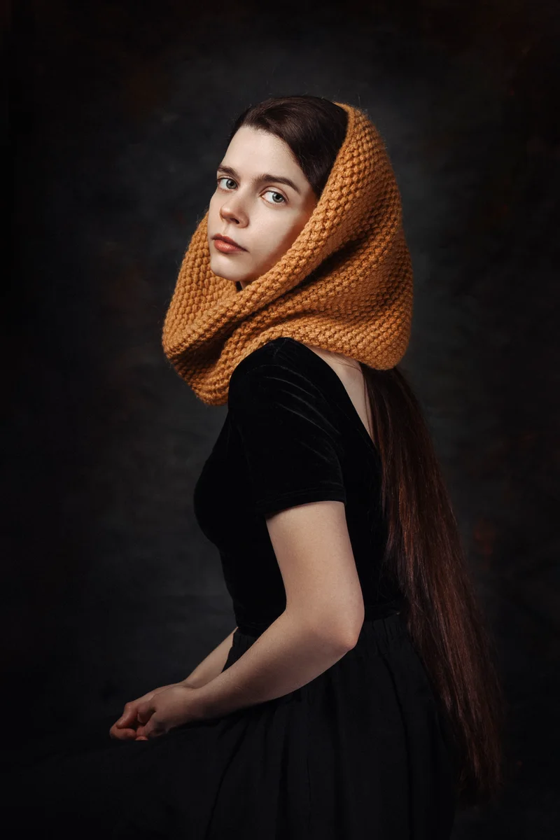 Woman with an orange knitted snood over her head and a black velvet dress in painterly light